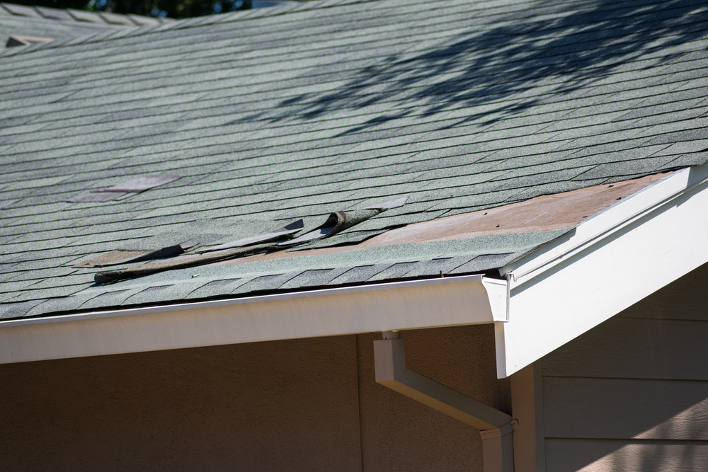 Asphalt shingle roofing on a residential home with visible damage, curled edges, and exposed sheathing. Close up